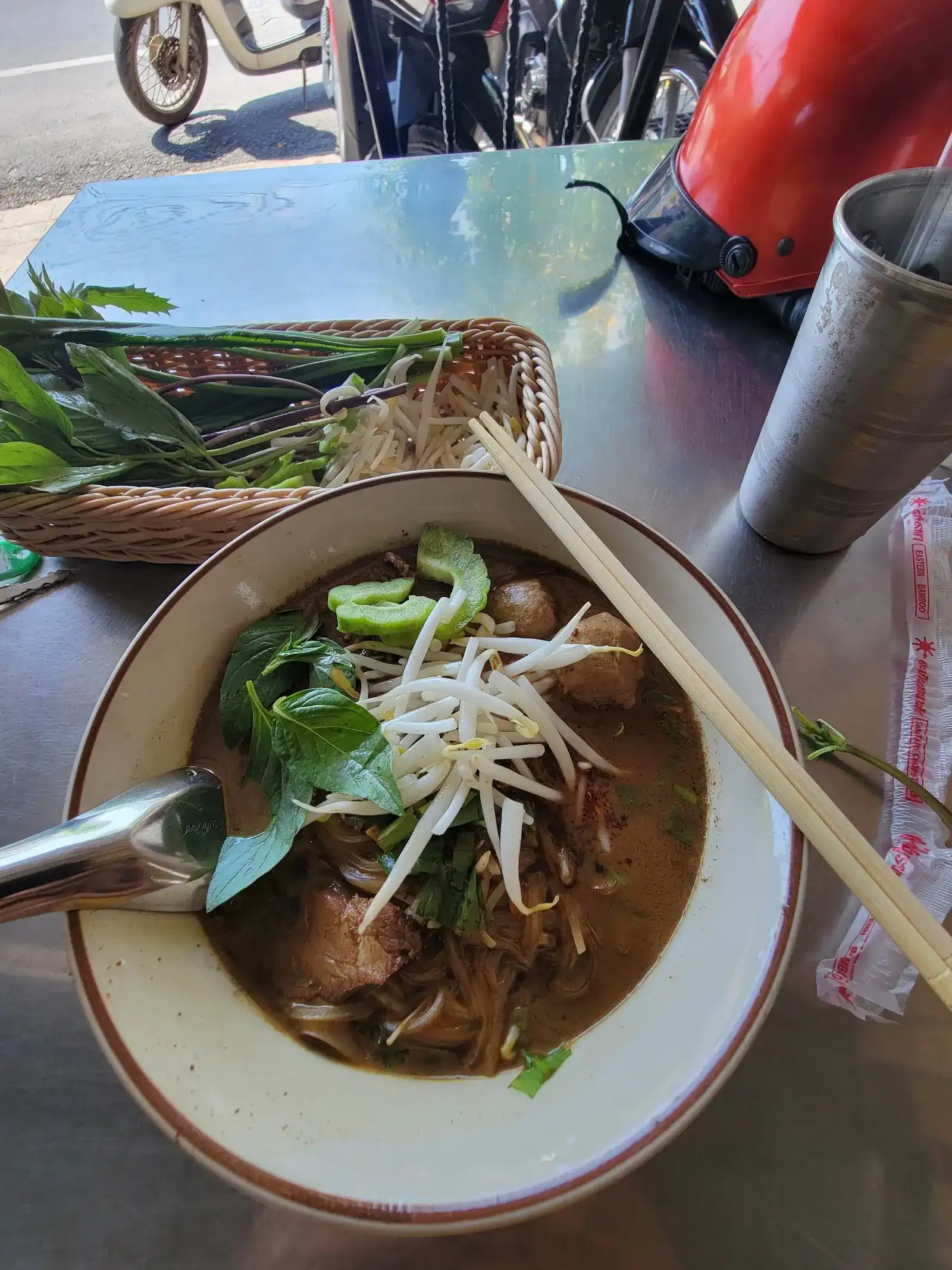 A bowl of boat noodles with beef, meatballs, Thai basil and bean sprouts, a basket of fresh herbs behind it on a steel table