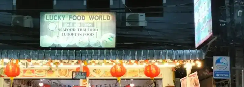 The front of the restaurant at night: the lit sign, a row of lights and red lanterns over the open dining room / หน้าร้านยามค่ำ ป้ายไฟ ไฟประดับ และโคมสีแดงเหนือห้องอาหารที่เปิดโล่ง
