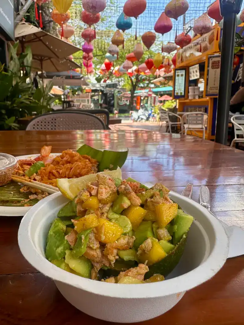 Grilled avocado with mango in a bowl, a pad thai behind it and paper lanterns overhead