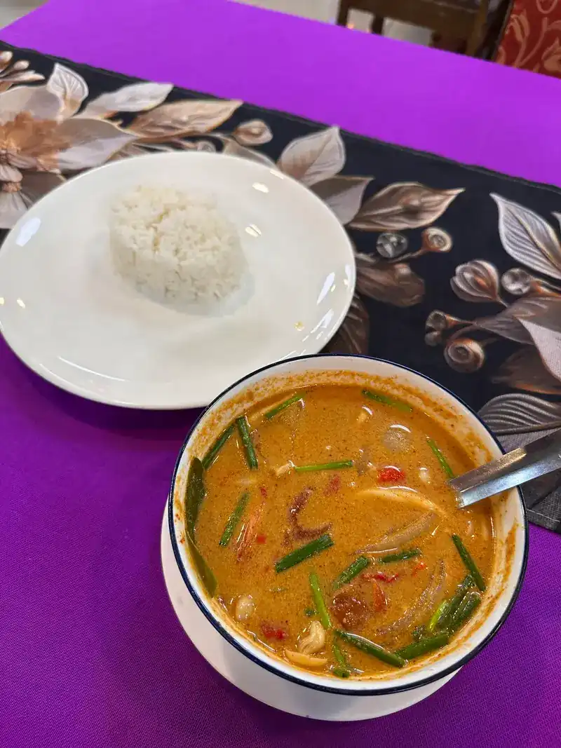 A bowl of spicy orange soup with lemongrass and a plate of rice on a purple tablecloth