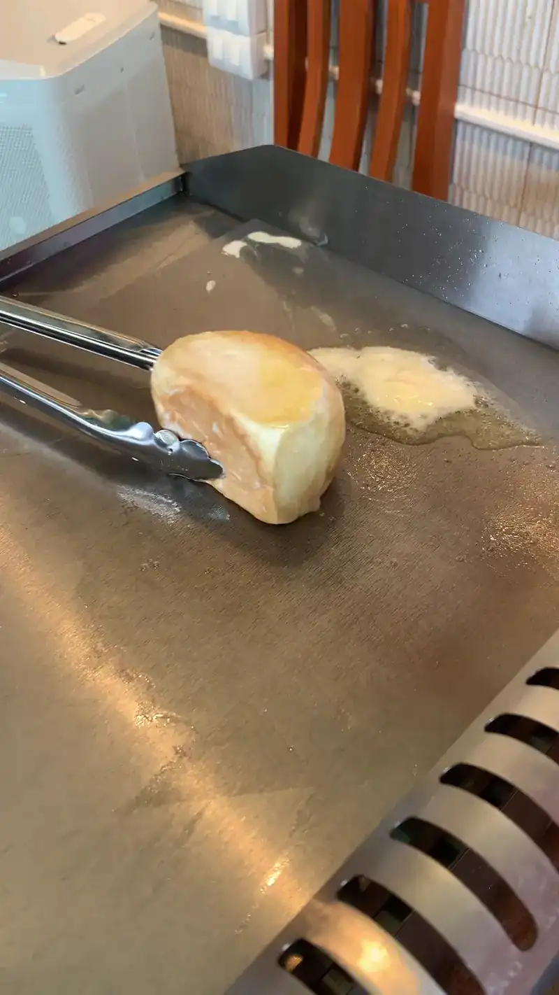 A bun being turned with tongs on a steel hot plate, butter melting beside it