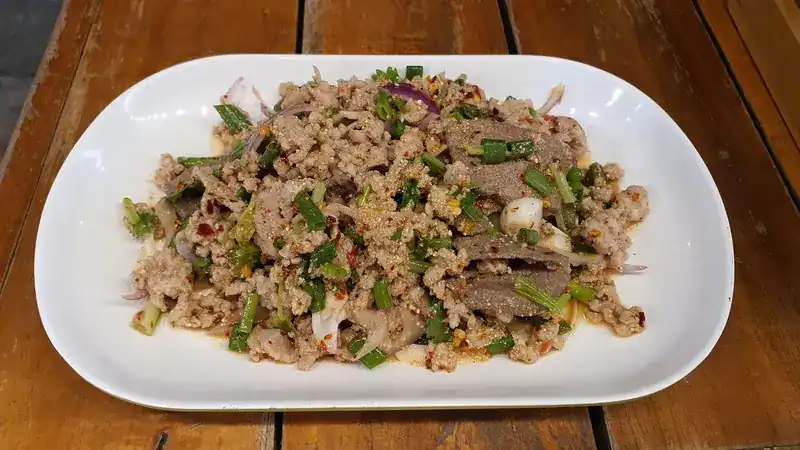 Minced larb with sliced meat, spring onion and chilli flakes on a white plate, on a wooden table