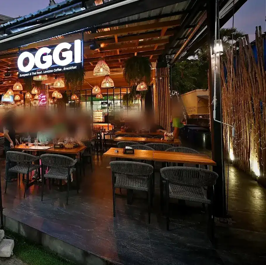The lit OGGI sign and woven rattan lamps over the open terrace at dusk / ป้ายไฟ OGGI และโคมไฟหวายสานเหนือระเบียงร้านยามพลบค่ำ