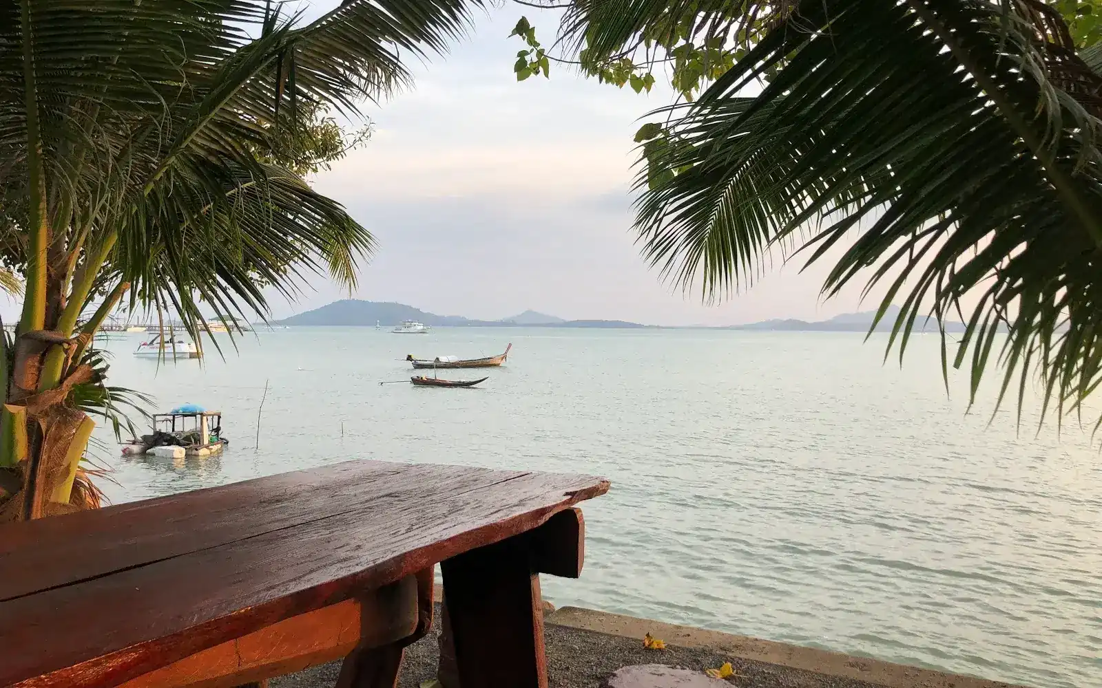 An empty timber table under palm trees, looking out over the bay with longtail boats and islands