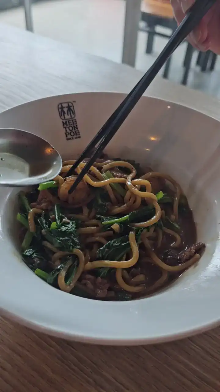 Chopsticks lifting yellow noodles and greens from a dark sauce in a Mee Ton Poe bowl