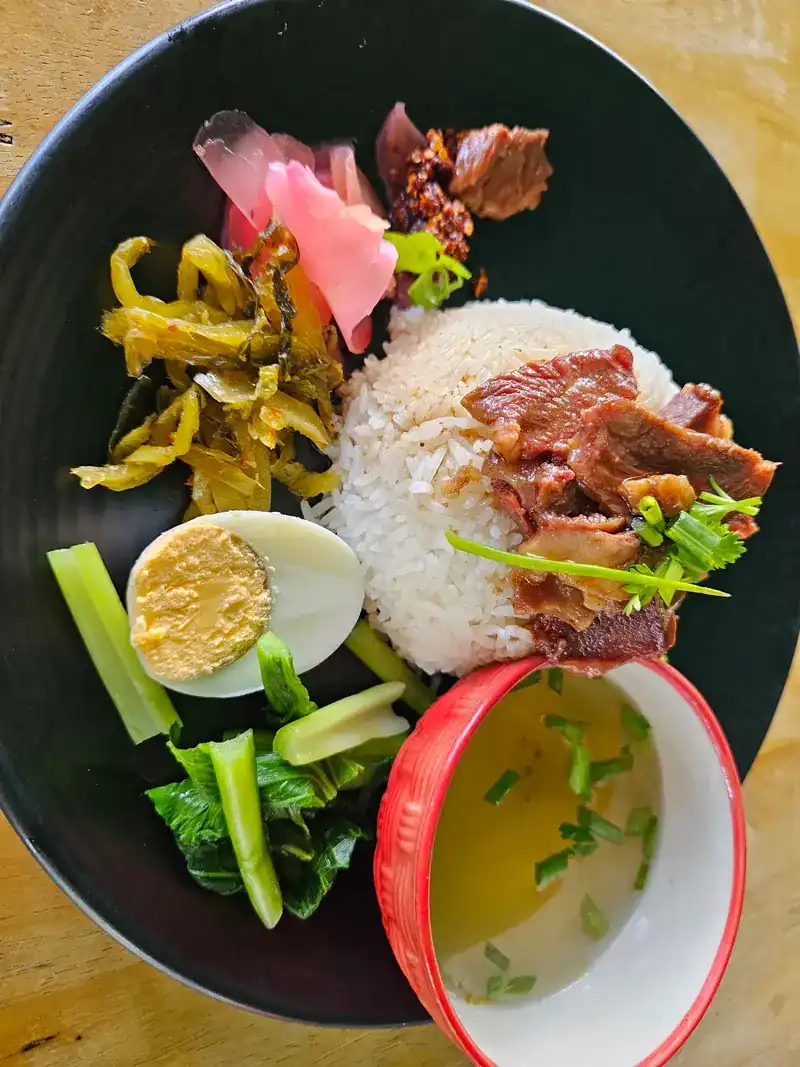 Rice with slices of stewed beef, half a boiled egg, pickled mustard greens, greens and a small cup of clear soup on a black plate