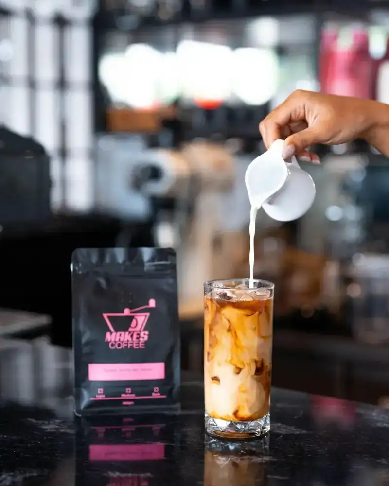 Milk poured into an iced coffee beside a bag of Makes Coffee