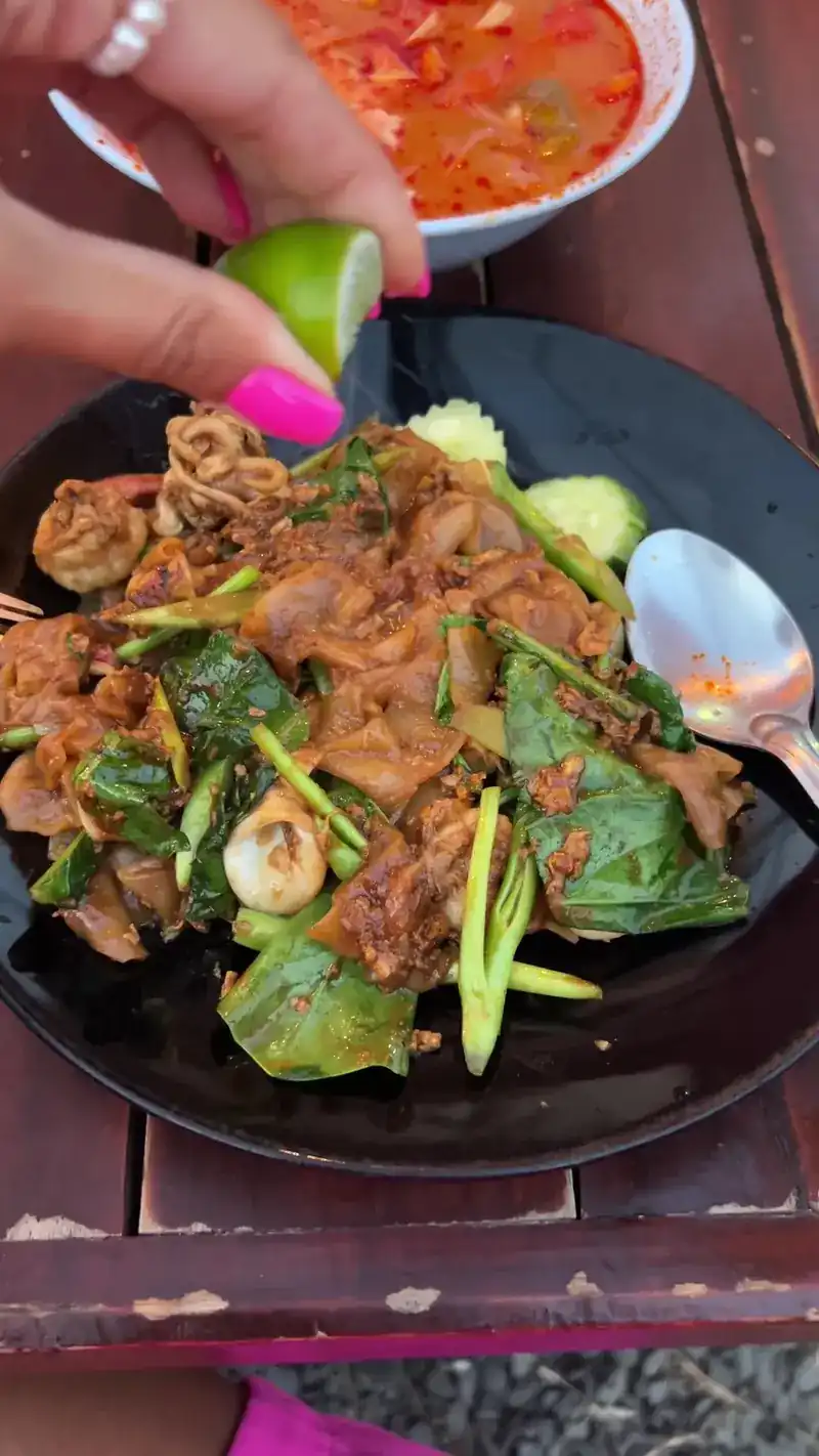 Wide rice noodles stir-fried with squid and greens, a hand squeezing lime over the plate