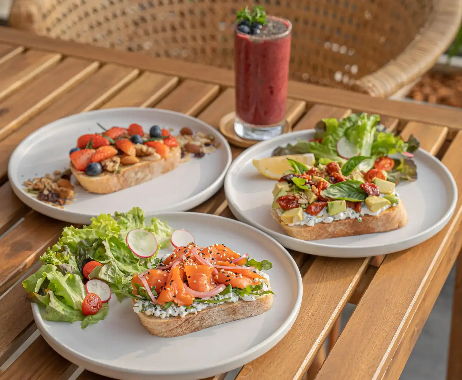 Three open toasts on white plates, topped with salmon, avocado and berries, with a berry smoothie on a wooden table