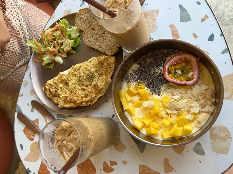 An omelette with salad and bread beside a smoothie bowl of mango, chia, almonds and passion fruit, with iced coffee