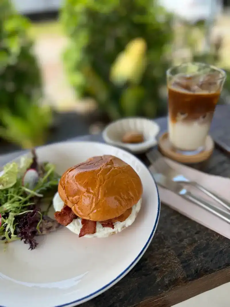 A brioche bun filled with bacon, with salad leaves and an iced coffee behind