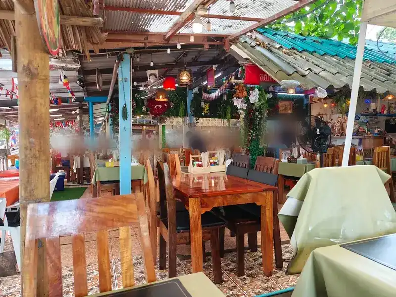 The open-air dining room with wooden tables, fans, lanterns and a wall of flowers