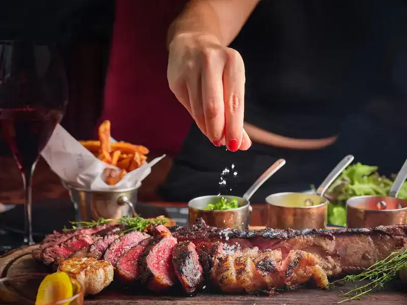 Sliced grilled steak on a wooden board with small copper pots of sauce, a hand adding a last pinch of salt
