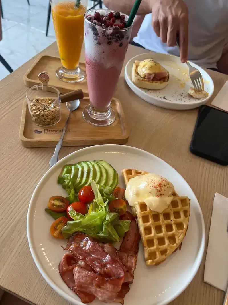 A waffle topped with a poached egg and hollandaise, with bacon, avocado and salad, next to a berry drink