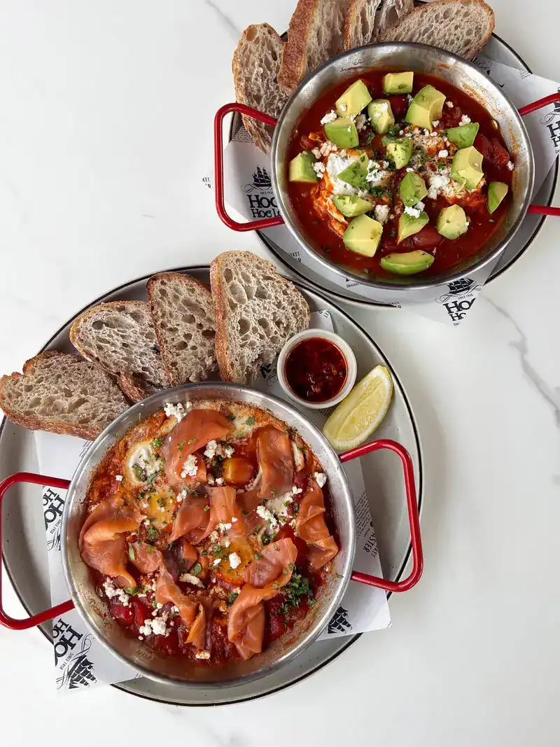 Two pans of eggs baked in tomato sauce, one topped with avocado and one with smoked salmon, with slices of sourdough