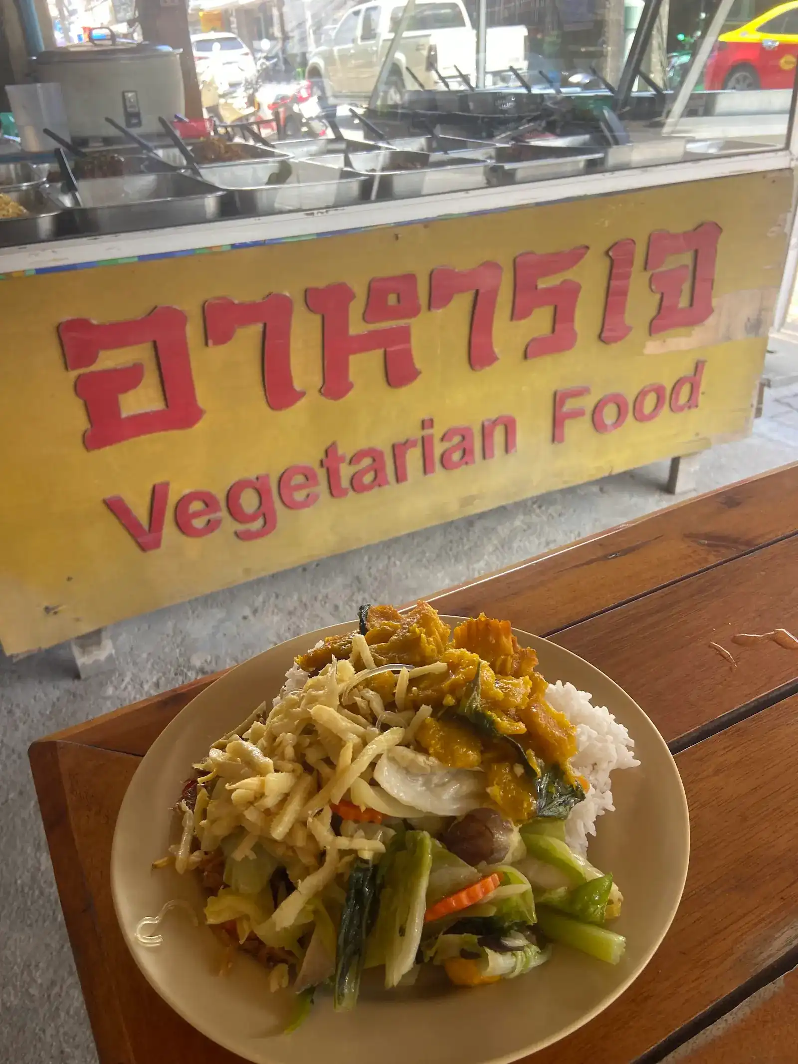A plate of rice with stir-fried vegetables in front of the shop's yellow sign, อาหารเจ Vegetarian Food