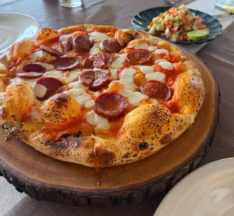 A pizza with sliced sausage and mozzarella on a wooden board, a small salad with lime behind it