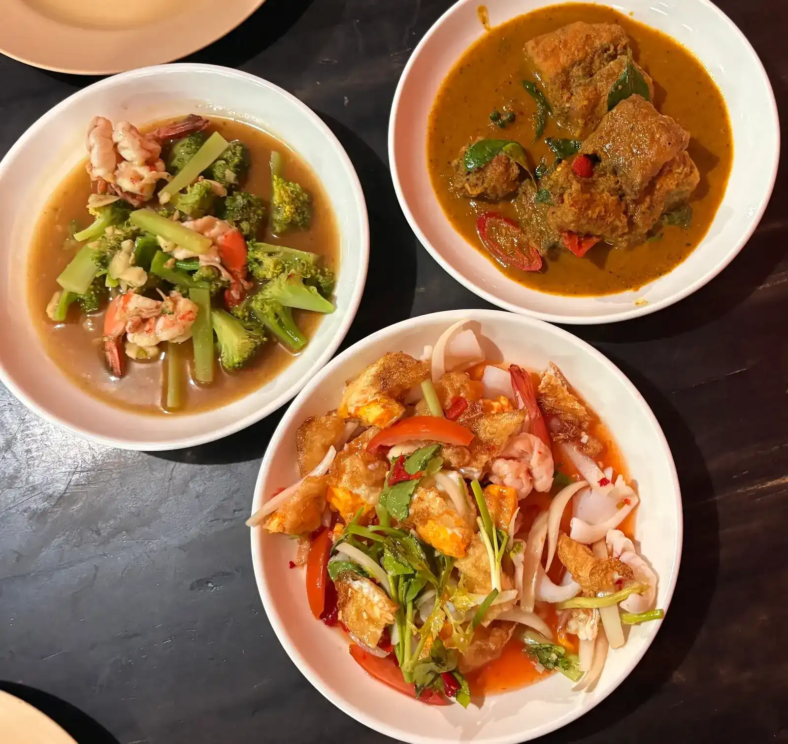 Three dishes on a dark table: prawns with broccoli, a thick curry, and a spicy salad of fried egg, squid and prawns