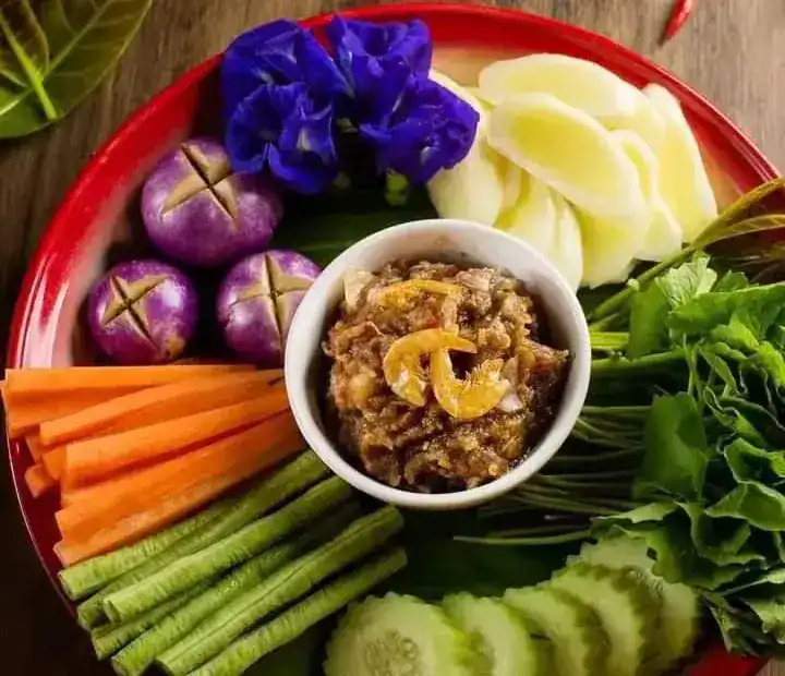 A chilli dip in a small bowl, ringed with fresh vegetables, round eggplants and butterfly pea flowers