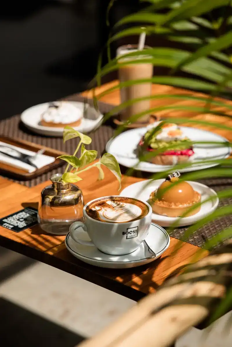 A latte in a BOOST cup beside a caramel tart and an avocado toast on a sunlit table