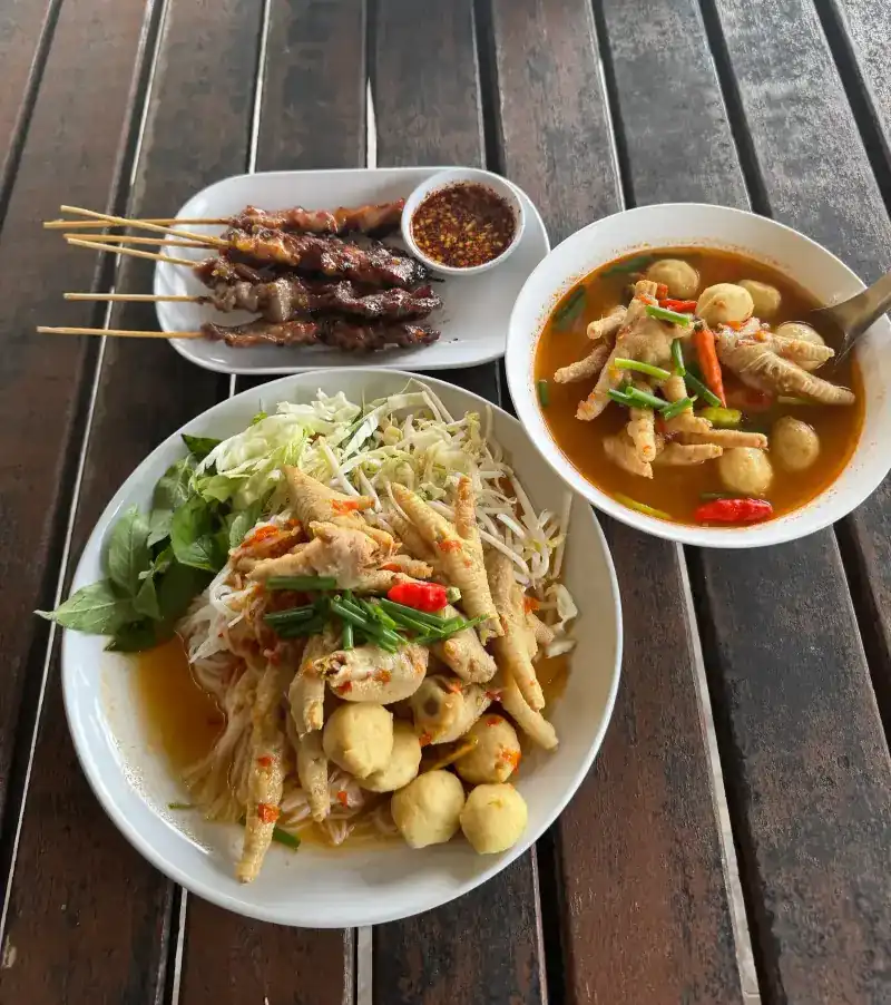 Grilled skewers with chilli dip, rice noodles with chicken feet, and chicken-feet soup on a wooden table / ไม้ย่างกับน้ำจิ้ม ขนมจีนตีนไก่ และต้มตีนไก่ บนโต๊ะไม้