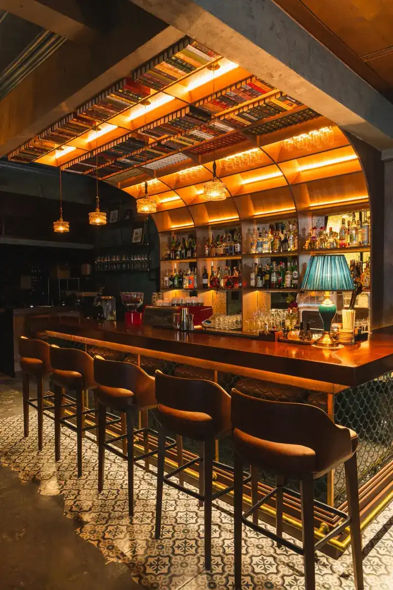 The bar: a long wooden counter with velvet stools under a vaulted, amber-lit back shelf