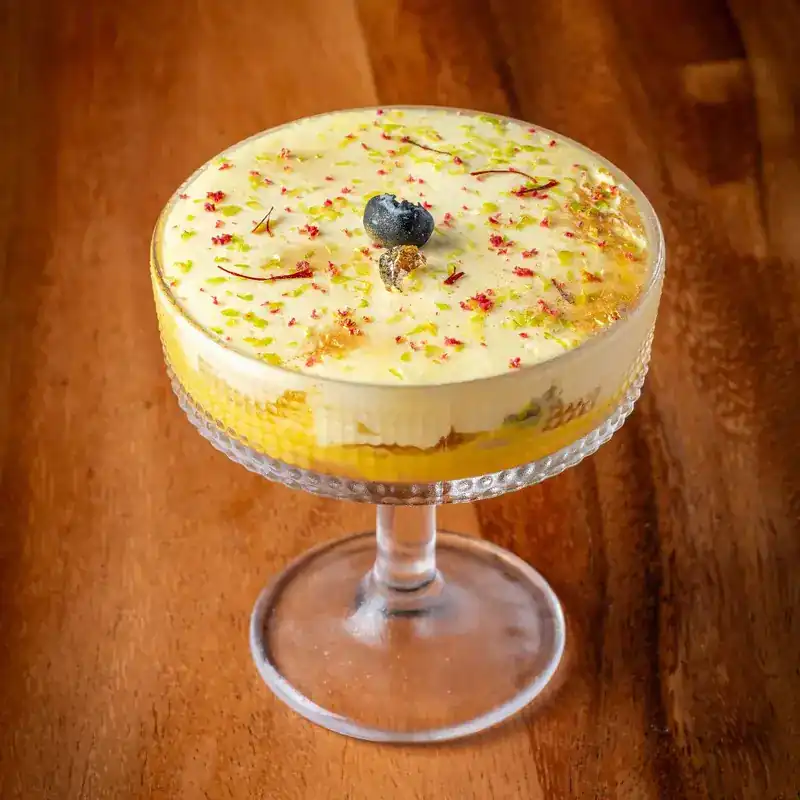 A layered Indian dessert in a glass coupe, with saffron threads and a blueberry on top