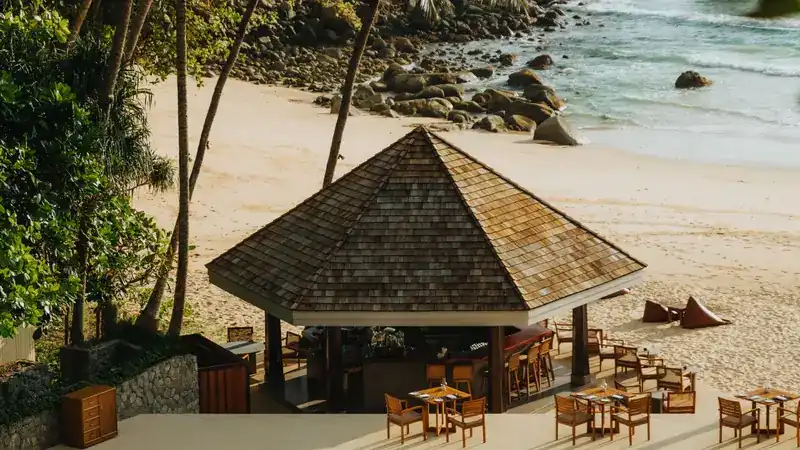 The Beach Bar pavilion with a shingled roof, tables on the deck and the sand and rocks of the bay behind