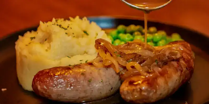 Two sausages on mashed potato with peas, onion gravy being poured over