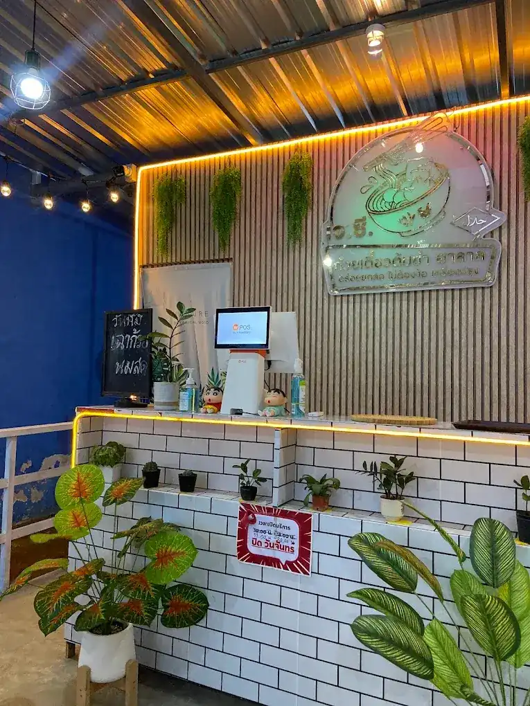 เคาน์เตอร์ร้านเจซี กระเบื้องขาว ไฟสีทอง ต้นไม้ และป้ายร้านบนผนังไม้ระแนง