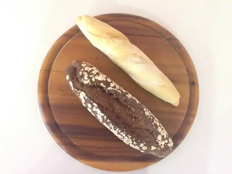 Two baguettes on a round wooden board, one white and one dark with oats