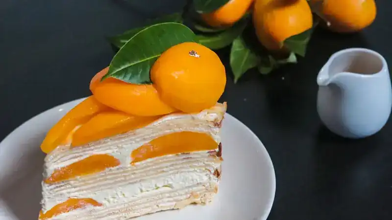 A slice of crepe cake layered with cream and orange fruit on a white plate