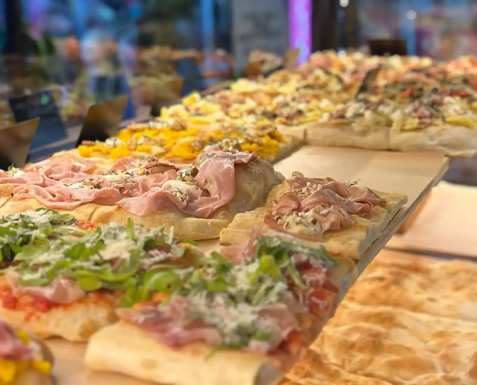 Slices of pizza alla pala lined up on wooden boards at the counter