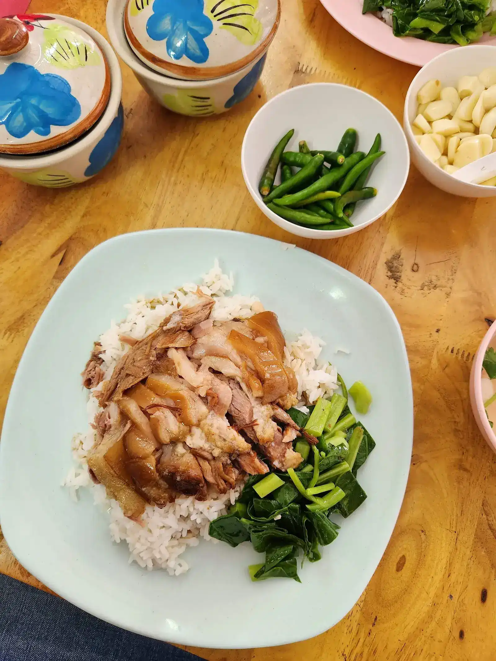 Pork leg rice with blanched greens, beside bowls of green chillies and raw garlic and two hand-painted lidded jars
