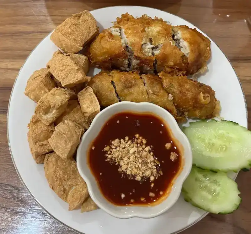 Fried rolls cut into pieces on a plate with fried tofu, cucumber and a dipping sauce topped with crushed peanuts