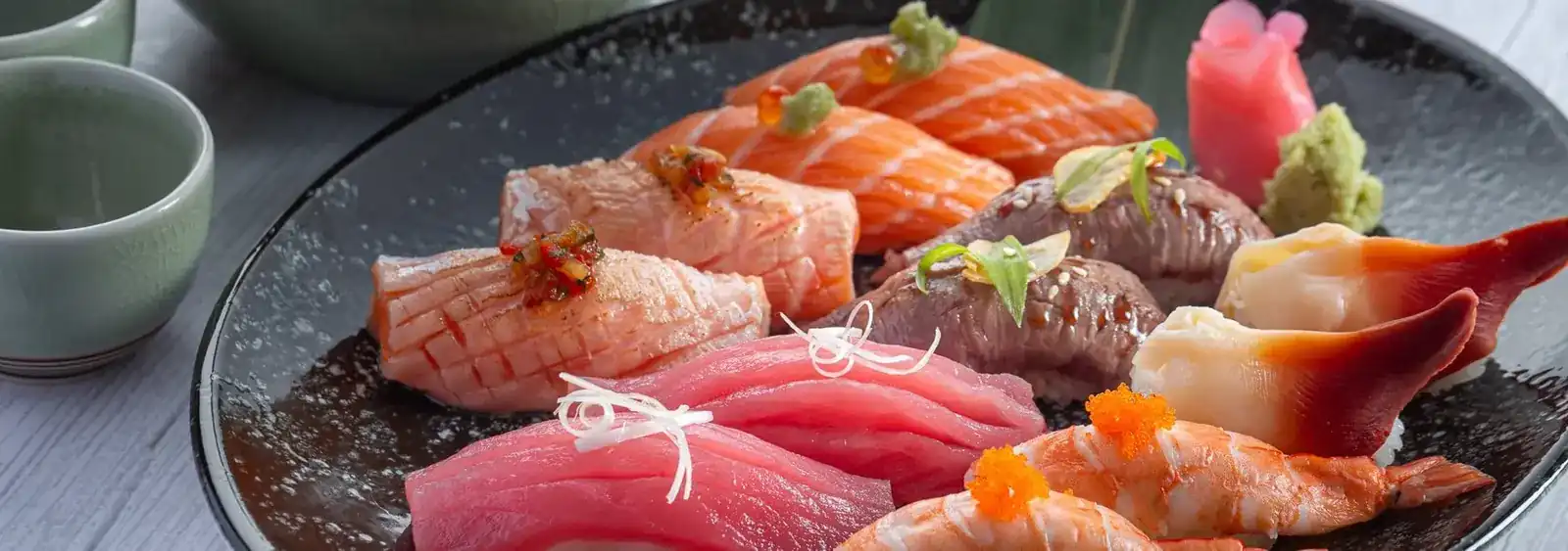Nigiri sushi on a dark plate, with celadon tea cups
