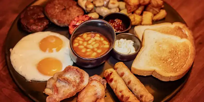 A full breakfast plate with fried eggs, sausages, bacon, baked beans, potatoes and toast