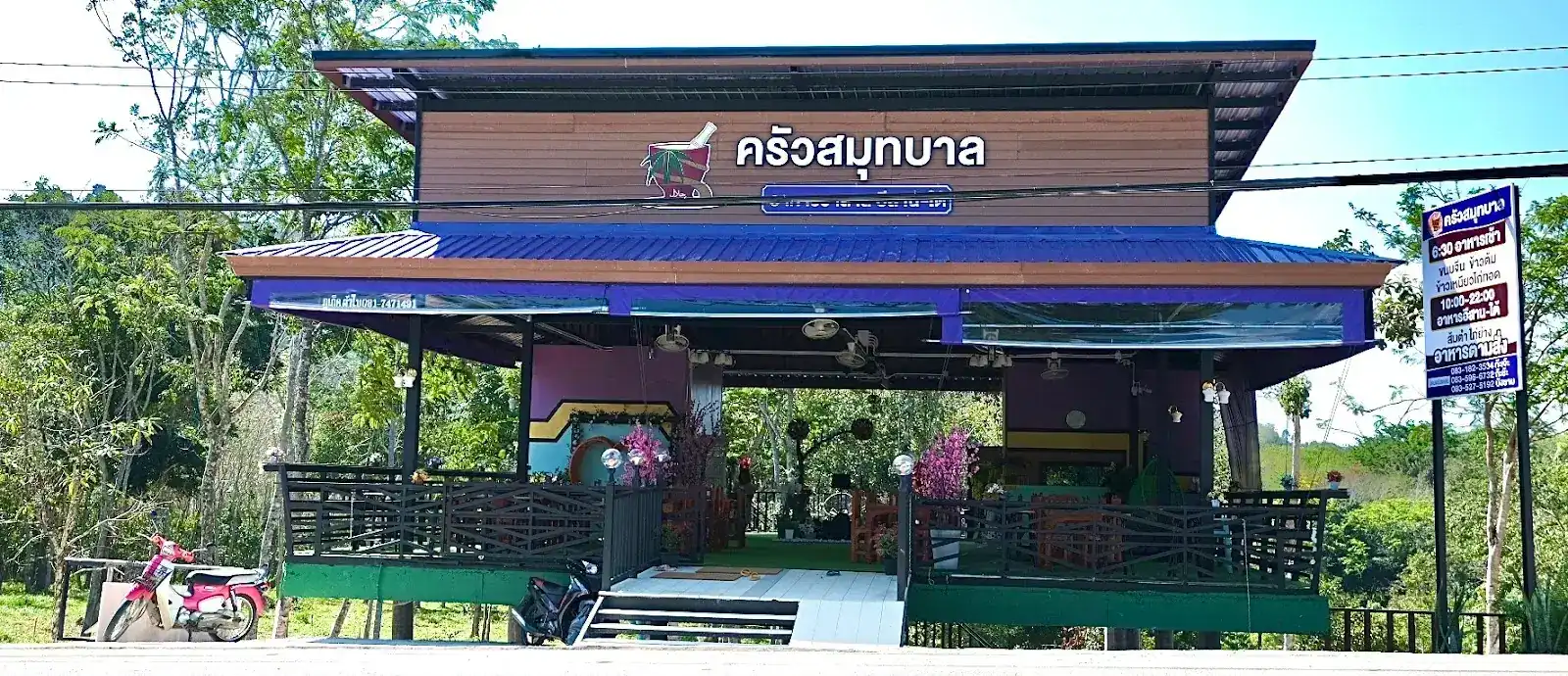 The front of Krua Samutbal seen from across the road: a wooden pavilion with a blue roof and the name sign in Thai