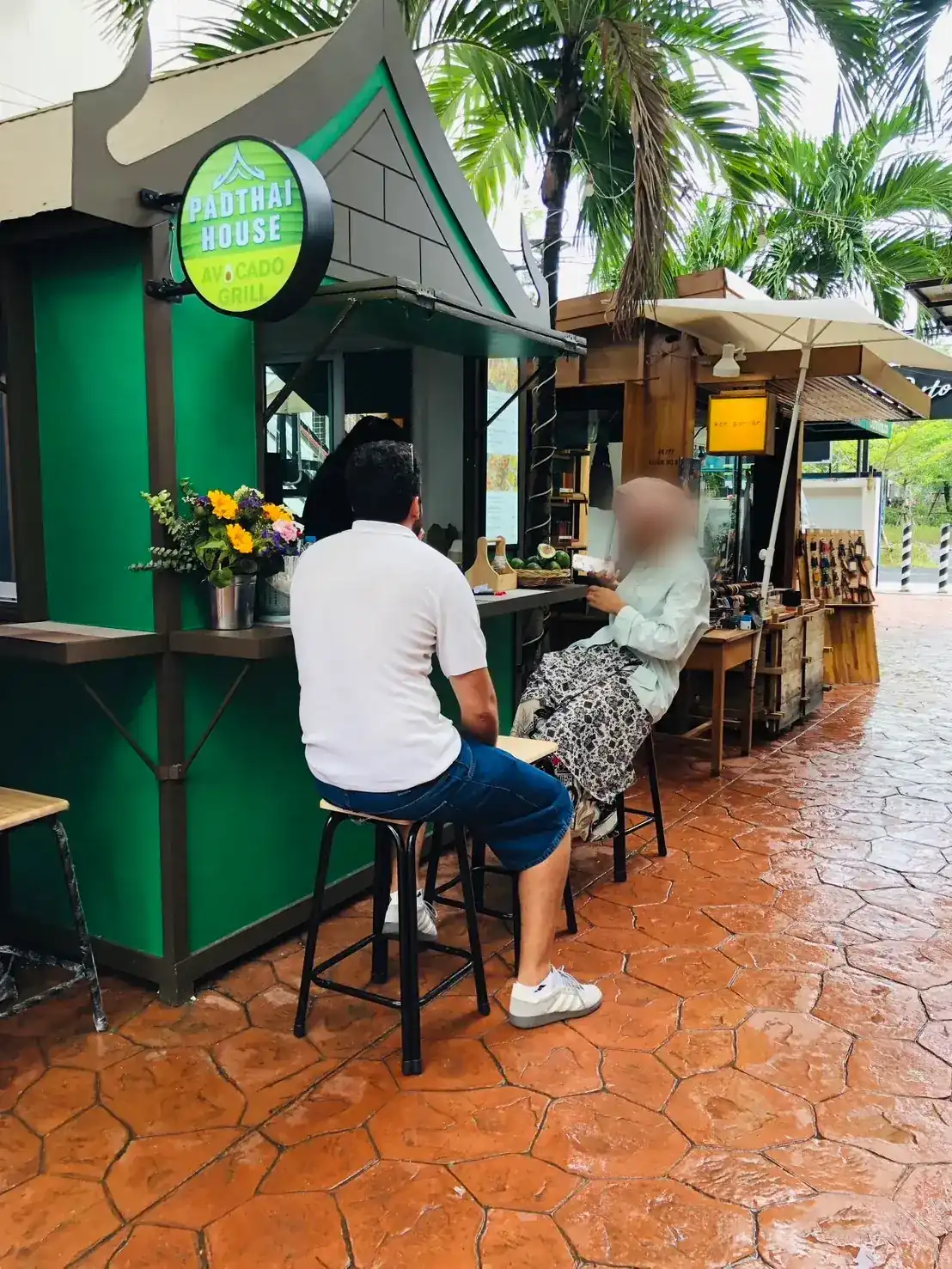 The green Padthai House kiosk with its round sign and counter stools, under palm trees
