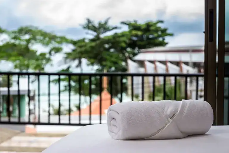 A rolled white towel on a bed, the balcony rail and trees behind it