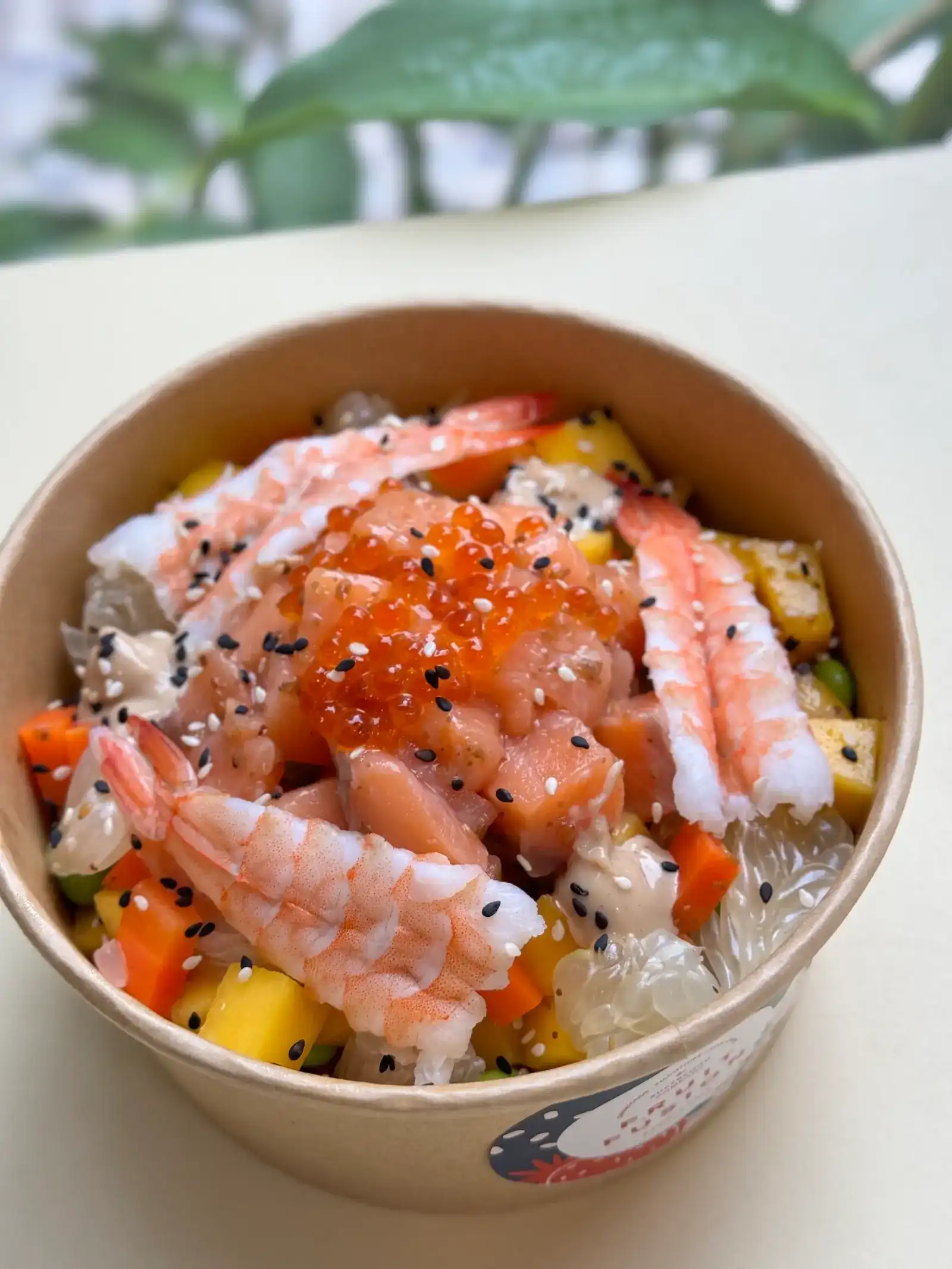 Poke bowl in a paper bowl, topped with shrimp, salmon roe, mango and black sesame