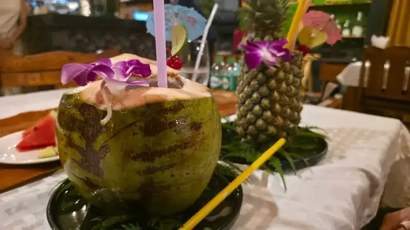 Two drinks dressed with orchids and paper umbrellas, one in a young coconut and one in a pineapple