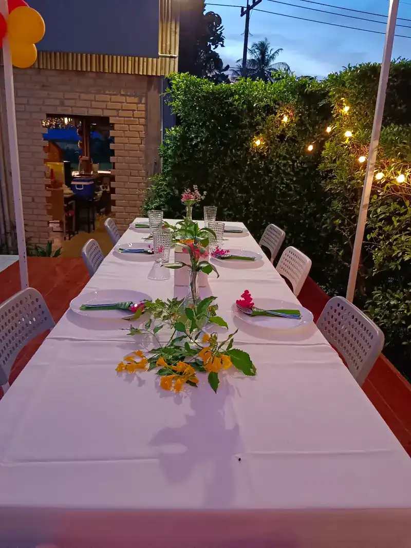 A long table outdoors at dusk, laid with white plates and flowers, string lights in the hedge