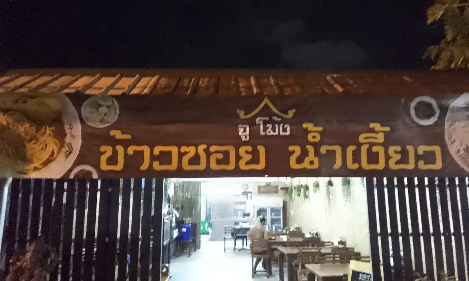 The shopfront at night: a dark wooden board with gold letters reading khao soi nam ngiao over the open door, wooden tables inside