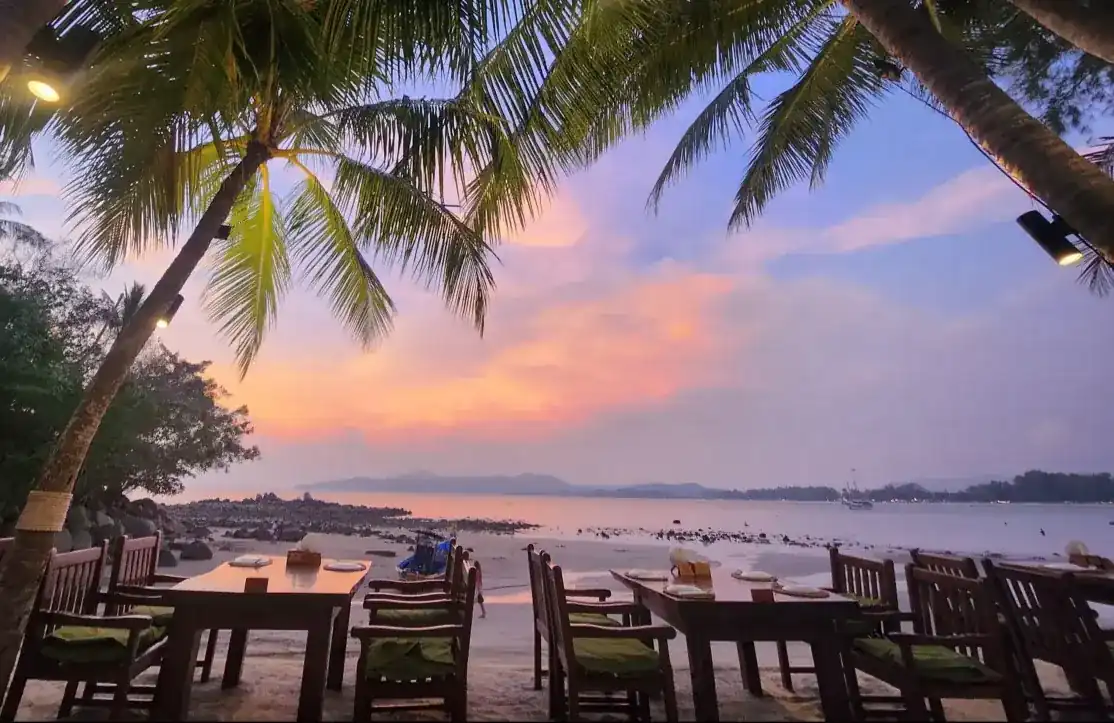 Tables on the sand under coconut palms at sunset, the sky and sea glowing pink and orange