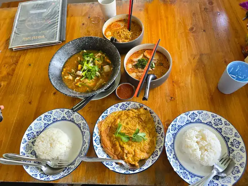 A wooden table set with noodle soups, a Thai omelette and plates of rice