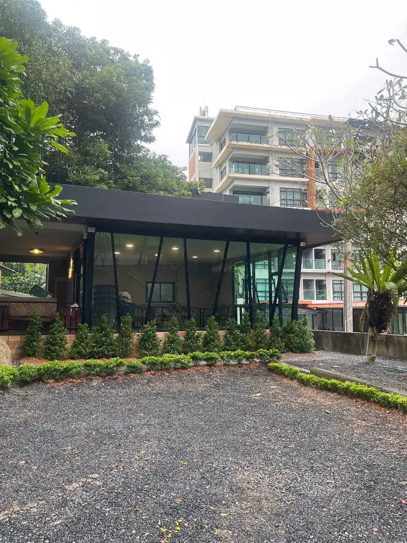 The restaurant's glass pavilion with a dark roof, set behind a low hedge and a gravel yard under tall trees