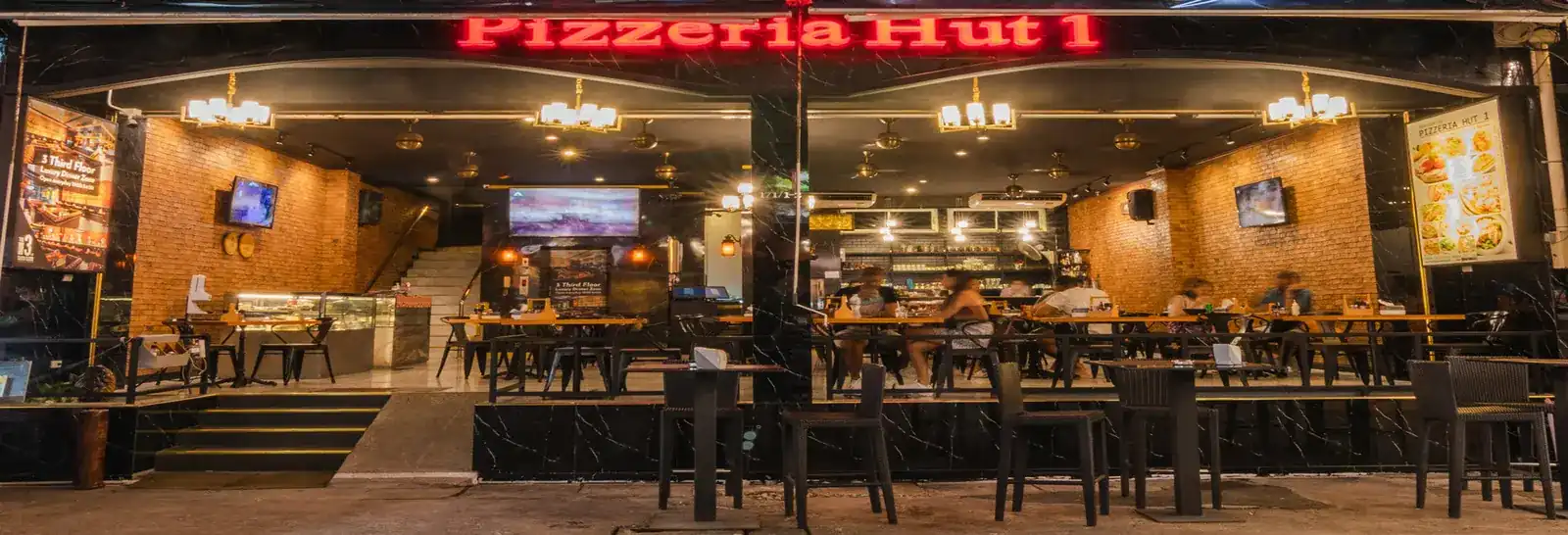 The open front of Pizzeria Hut 1 at night, its red neon sign above brick walls and wooden tables