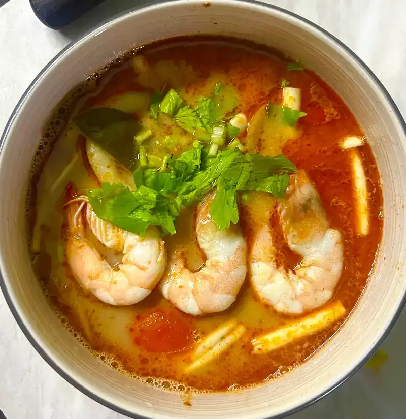 A bowl of red soup with prawns, tomato and herbs