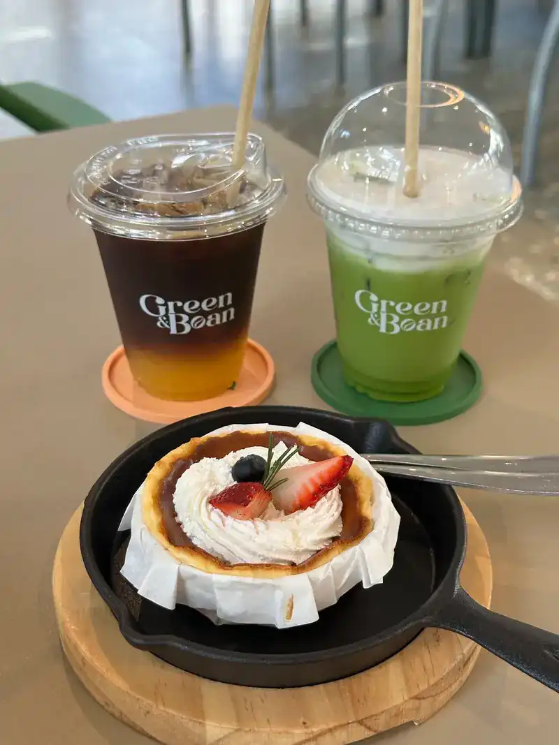 Two iced drinks in Green & Bean cups, one coffee and one green, beside a cake topped with cream and berries in a small iron pan